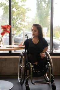 A South Asian person in a wheelchair looks thoughtfully to the side while taking notes. She is backlit by the window she's in front of and wearing a teal shirt with black pants and shoes.