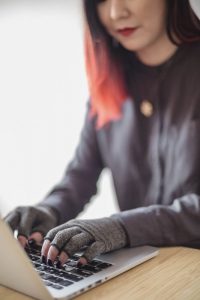An Asian disabled woman types on a laptop while wearing compression gloves. The hands and keyboard are the focal point.