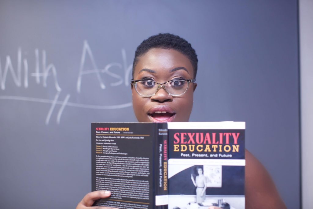 image of Ashley Cobb in front of chalkboard with "with ashley" written on it. Ashley is holding a sexuality education book and looking into the camera