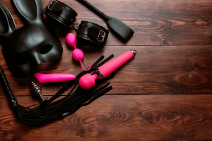 BDSM mask, crop, flogger and cuffs next to pink ben wa balls, wand massager, and small vibrator -- when used together would make a hot story about sex as part of a sex toy review