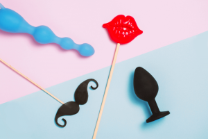 Blue anal beads and a black butt plug next to a toy moustache on a stick and toy red lips on a stick that looks like a role play situation which may be great for writing a sex toy review
